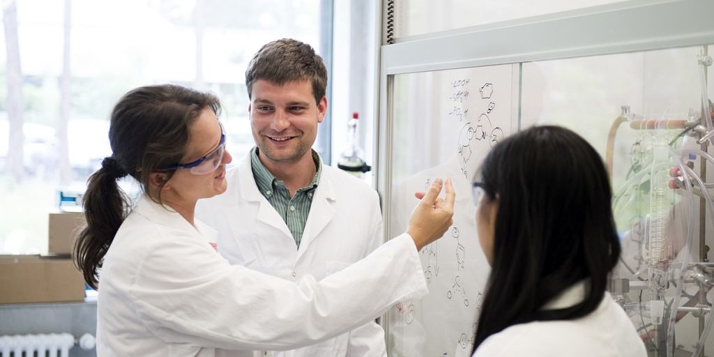 Two female researchers with long brown hair and on male researcher with short brown hair, all wearing laboratory cloaks, discussiing chemical research schemes on a transparent modern blackboard