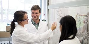 Two female researchers with long brown hair and on male researcher with short brown hair, all wearing laboratory cloaks, discussiing chemical research schemes on a transparent modern blackboard