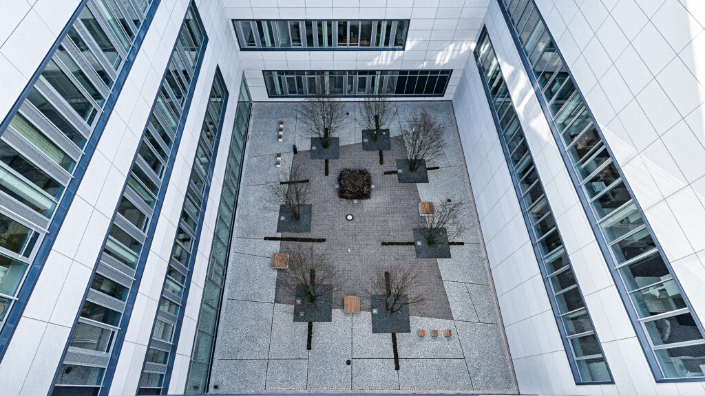 Viwe of the inner yard of the IZNF research building - white walls on three sides, 4 stirages high, with several window rows. In the middle is a grey ston floor wir some small trees