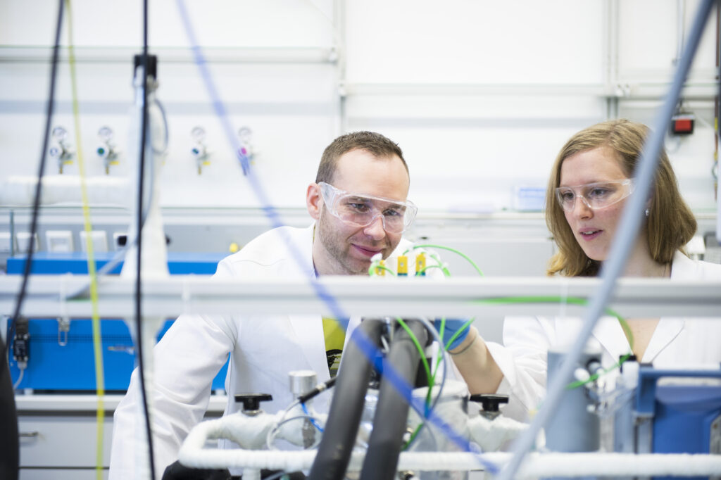 A female and a male scientist, both wearing lab coats and lab glasses, working together on a machinery at a chemical reaction laboratory