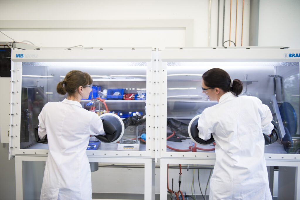 Two female scientists with long brown hair wearing white lap coats - both seen from the back. They have their hand in a glovebox (scientivic instrumentation) and handing a bottle with chemicals to each other, protected by the big black gloves of the glove box